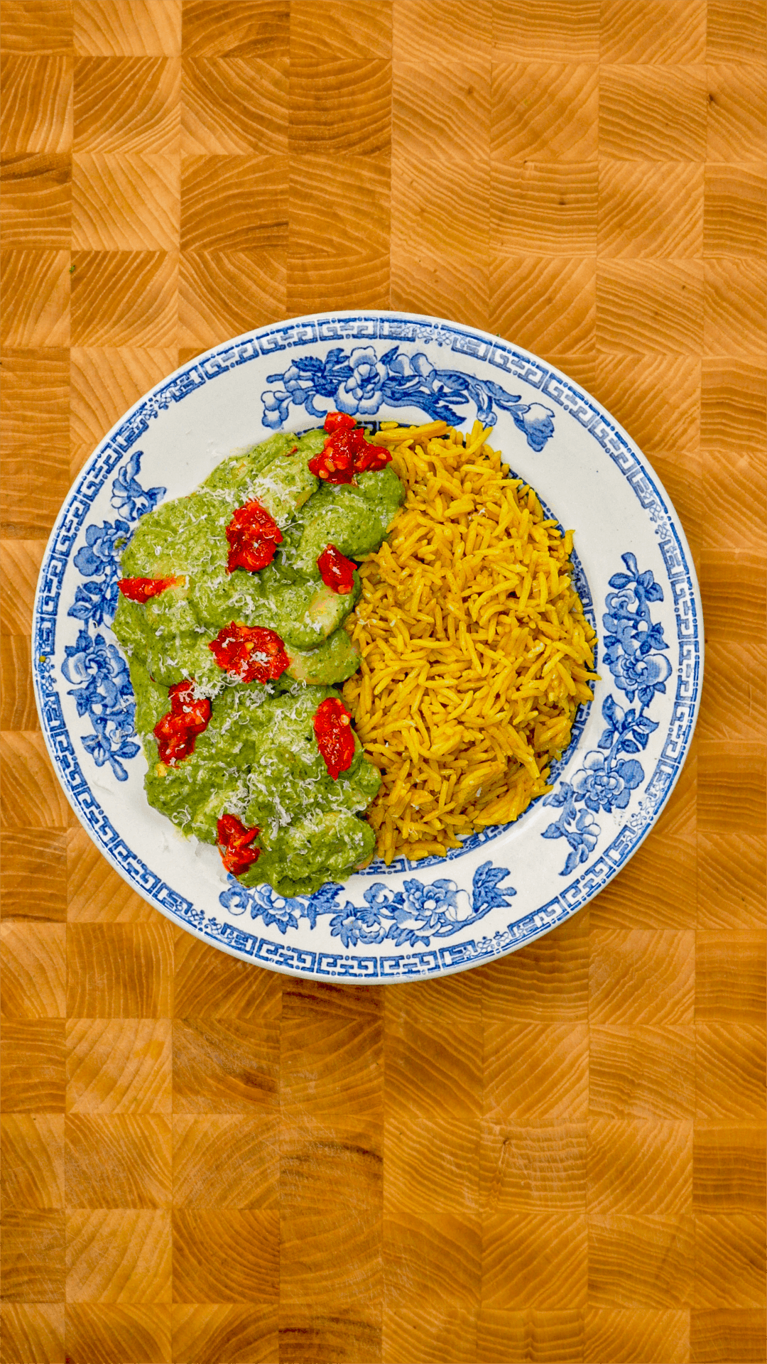 Overhead view of Creamy Zucchini Butter Beans served on a blue and white floral plate, with vibrant green zucchini butter bean sauce topped with sun-dried tomatoes and grated cheese alongside Royal Yellow Basmati Rice, on a wooden checkerboard surface