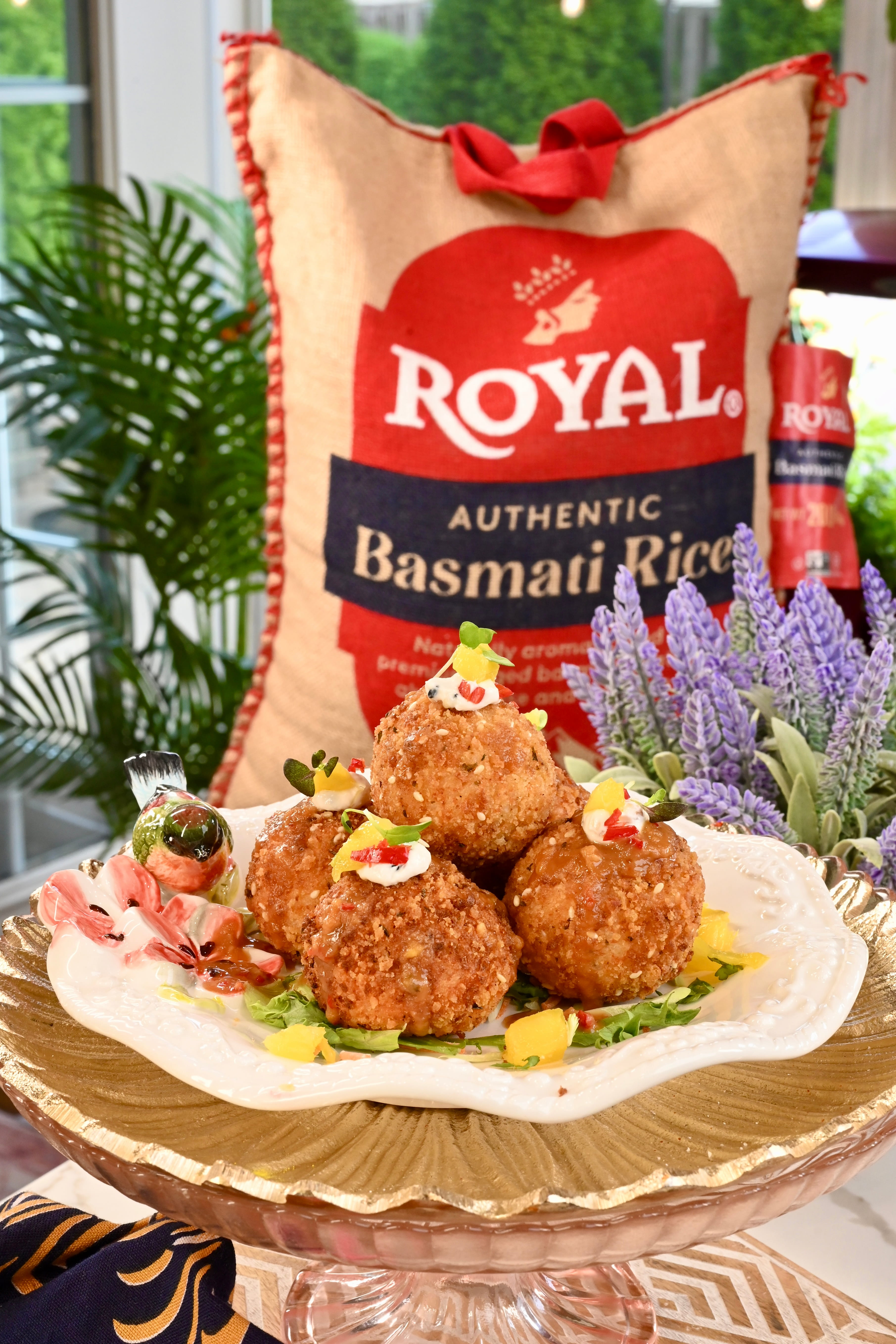 Crispy golden Spicy Mango Coconut Bites piled on an ornate white and gold serving platter, garnished with lemon aioli, microgreens, and chopped red chilies over a spring salad with mango pieces, with a bag of Authentic Royal Basmati Rice in the background