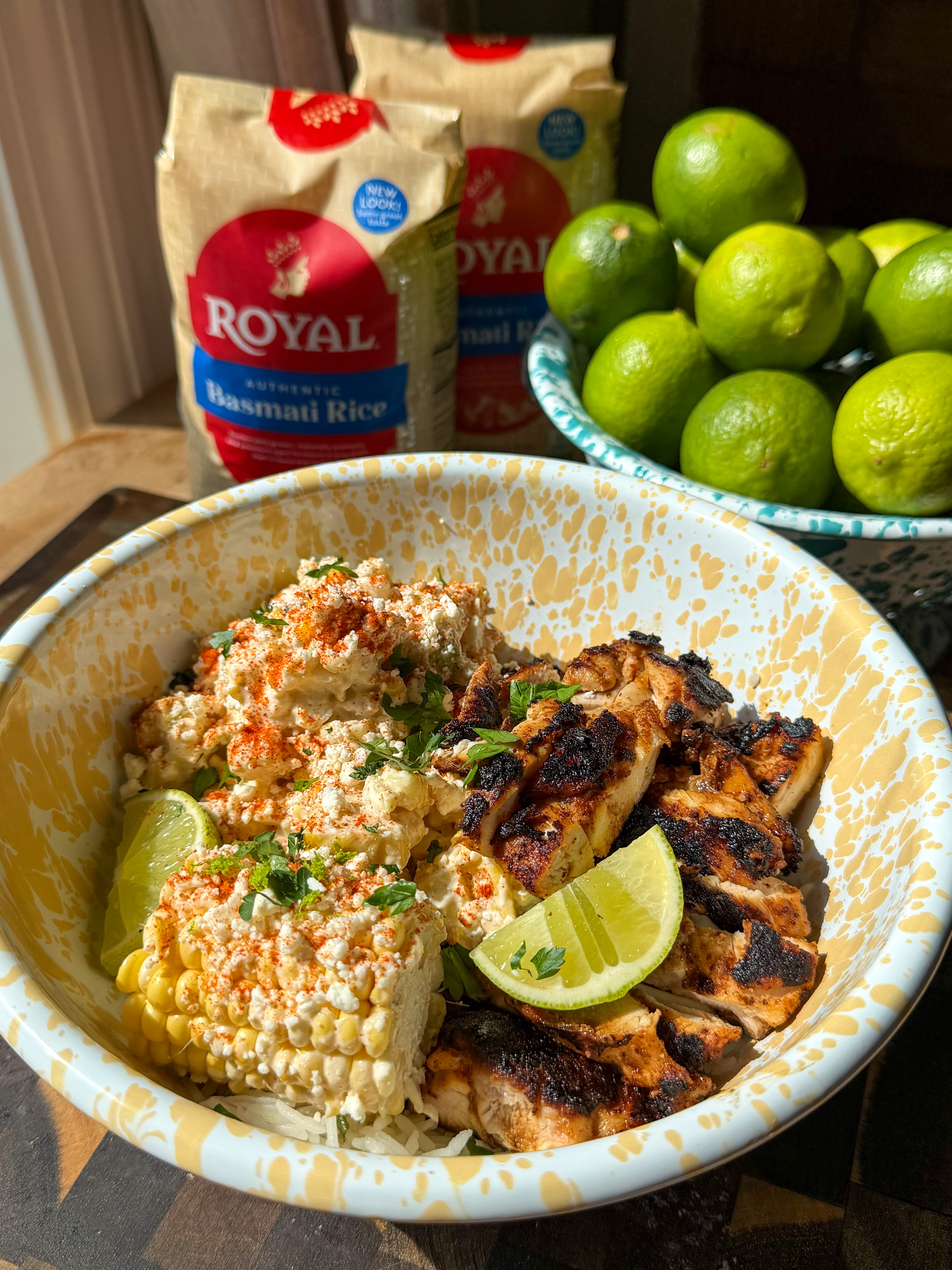 A yellow and white speckled bowl filled with Mexican Street Corn Chicken and Rice, featuring charred sliced chicken, creamy street corn topped with crumbled feta, chili powder, and fresh cilantro, served over Royal Authentic Basmati cilantro lime rice with lime wedges, with two bags of Royal Authentic Basmati Rice and a bowl of fresh limes in the background