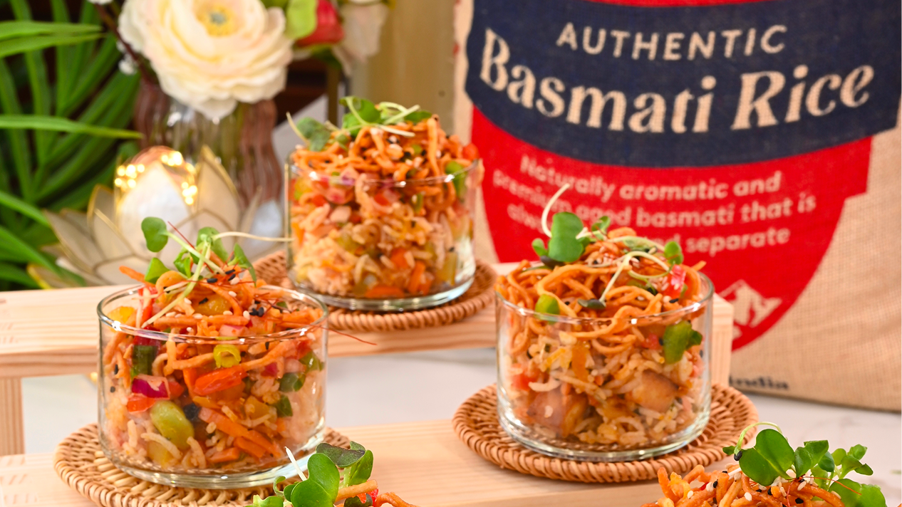 Crunchy Chinese Rice Bhel Cups served in clear glass cups, layered with Schezwan fried basmati rice, colorful vegetables, crispy noodles, and fresh microgreens, arranged on woven coasters with a bag of Royal Authentic Basmati Rice in the background.