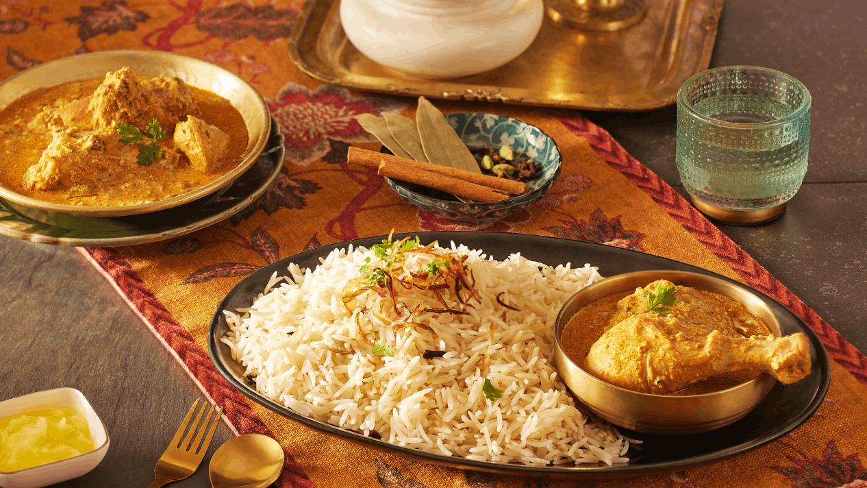 Fluffy Royal® Chef's Secret™ Basmati Rice served with creamy Awadhi Chicken Korma in a golden bowl on a dark platter, with whole spices and an embroidered table runner